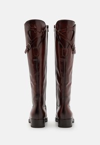 Botas altas de cuero marrón con una textura suave, que cuentan con un cierre de cremallera en la parte posterior, correas con hebilla y un tacón cuadrado robusto.