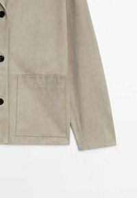 Vaaleanharmaa mokkajakku, jossa on suora leikkaus, mustat napit ja yksi läpätasku vasemmalla puolella. Pehmeä pinta, yksinkertainen muotoilu.