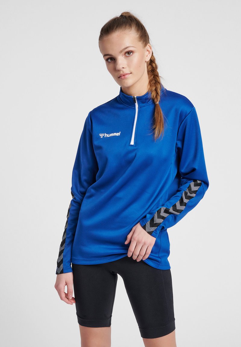 Maglia sportiva a maniche lunghe blu con colletto a mezzo zip, caratterizzata da accentuazioni nere a motivo chevron sulle maniche. Indossata con pantaloni corti neri aderenti.