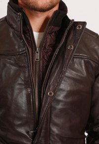 Braune Lederjacke mit Stehkragen, Reißverschluss mit Druckknöpfen und wattierter Innenauskleidung. Glatte Textur mit sichtbaren Nähten.