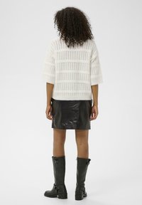 Femme aux cheveux bouclés portant un pull en tricot blanc, une mini-jupe en cuir noir et des bottes noires mi-mollet, se tenant de dos par rapport à l'appareil photo.