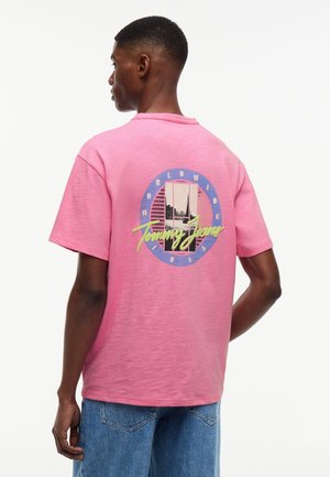 Άνδρας που φοράει ροζ μπλουζάκι Tommy Jeans με γραφικό λογότυπο και κείμενο σε μπλε και κίτρινο στο πίσω μέρος, σε συνδυασμό με μπλε τζιν παντελόνι.