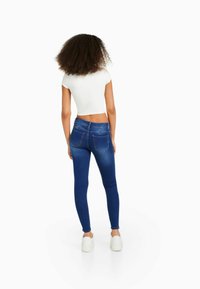 Jeans ajustados de mezclilla azul con cintura de altura media, con un desvanecimiento mínimo y bolsillos traseros. Combinados con una blusa blanca corta de manga corta.