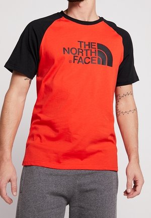 Mand iført rød og sort The North Face t-shirt med korte ærmer og grå bukser, der viser underarmstatoveringer på begge arme.