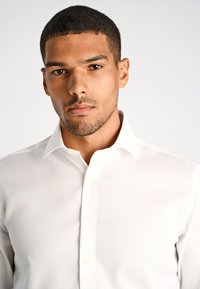 Camicia bianca con bottoni, colletto classico, texture liscia e sei bottoni sulla parte anteriore, indossata da un modello maschile su uno sfondo neutro.