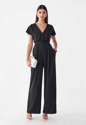 Donna che indossa una tuta nera a righe sottili con pantaloni larghi, scollo profondo a V e maniche corte, tiene in mano una pochette bianca texture e indossa sandali bianchi.