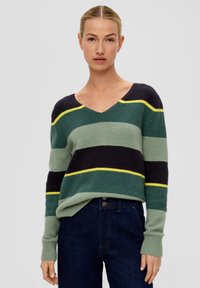 Gestreifter Pullover in Grün, Marineblau und Gelb. Mit V-Ausschnitt, gerippten Bündchen und weicher Textur. Kombiniert mit dunklen Jeans.