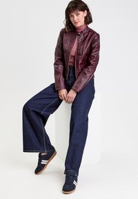 Giacca di pelle bordeaux con colletto alto, abbinata a jeans larghi scuri e sneakers blu navy con strisce bianche. Design pulito e moderno.