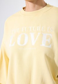 Persona vestida con un sudadera de color amarillo pálido y texto bordado en blanco que dice "EL FUTURO ES AMOR" en el pecho.