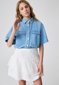 Lyseblå denimskjorte med knapper, der har to brystlommer og sølvhardware, kombineret med et lagdelt hvidt skørt med hulmønster.