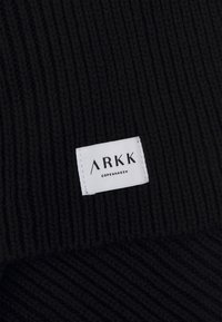 Caciulă tricotată neagră cu textură ribată și o etichetă rectangulară albă care afișează "ARKK COPENHAGEN" în text negru.