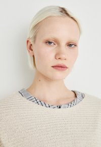 Beige kötött pulóver strukturált felülettel és kerek nyakkivágással, kék és fehér kockás ingnyakkal rétegezve.
