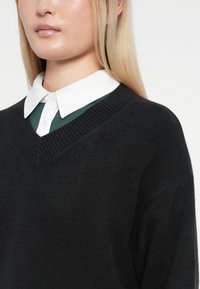 Fekete, texturált kötött pulóver V-nyakkal, zöld galléros ing fölött, fehér gallérral. Közeli felvétel a felső testről.