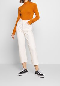 Oranges langärmeliges, tailliertes Oberteil mit hohem Kragen, kombiniert mit off-weißen, weit geschnittenen Hosen. Schwarze Sneakers mit weißen Akzenten vervollständigen das Outfit.