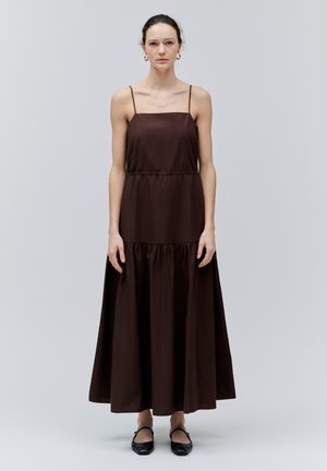 Kvinde med mørkt hår iført en lang brun ærmeløs kjole med tynde stropper og sorte flade sko, stående foran en ensfarvet grå baggrund.