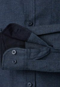 Close-up van een getextureerde donkerblauwe shirtmanchet met twee zwarte knopen, gedeeltelijk losgemaakt om de binnenste stofvoering te tonen.
