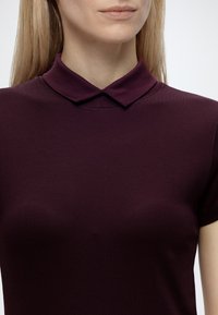 Femme aux cheveux blonds lisses portant un haut à manches courtes, col violet foncé, montrée des épaules au menton sur fond uni.