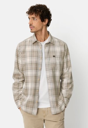 Beige ternet skjorte med knapper og lange ærmer, med hvide og blå linjer. Båret over en hvid T-shirt med rund hals. Lomme på venstre bryst.