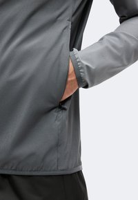 Main placée dans la poche latérale zippée d'une veste grise avec poignets élastiques, présentée sur un fond clair.
