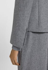 Primo piano di una persona che indossa un cappotto di lana grigio e una gonna abbinata, con i polsini a coste del maglione ben visibili e la mano rilassata al loro fianco.