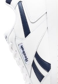 Baskets en cuir blanc avec des accents bleu marine, présentant des surfaces lisses et texturées, des coutures visibles et un logo sur le côté. Lacets à l'avant.