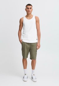 Hvid bomulds tanktop, olivenfarvede cargo shorts med sidelommer, og hvide sneakers med sorte striber; modellen viser en neutral holdning.
