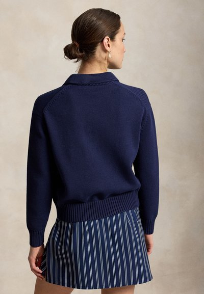 Femme avec les cheveux en chignon, vêtue d'un pull en tricot bleu marine et d'une jupe bleu marine à rayures verticales blanches, tournée dos à la caméra.
