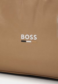 Tan spalvos rankinė su lygiu paviršiumi, ant priekio esantis prekės pavadinimas "BOSS" baltos, juodos ir auksinės spalvų akcentais.