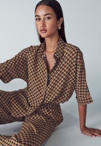 Brązowa i czarna koszula w wzory z geometrycznym wzorem, z kołnierzykiem i luźnymi rękawami, zestawiona z dopasowanymi luźnymi spodniami.