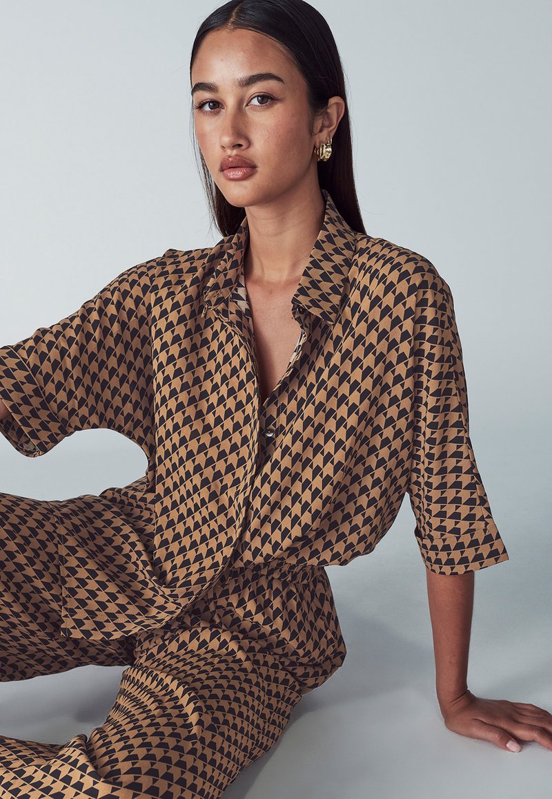 Brązowa i czarna koszula w wzory z geometrycznym wzorem, z kołnierzykiem i luźnymi rękawami, zestawiona z dopasowanymi luźnymi spodniami.