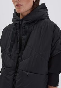 Gilet rembourré noir avec un col haut, une capuche et un motif matelassé en diagonale. Comprend deux poches latérales zippées et des cordons de serrage réglables.