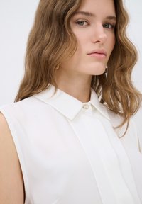 Witte mouwloze blouse met een klassieke kraag, verticale plooien en een kleine knoop bij de kraag. Glad, lichtgewicht stof met een zachte afwerking.