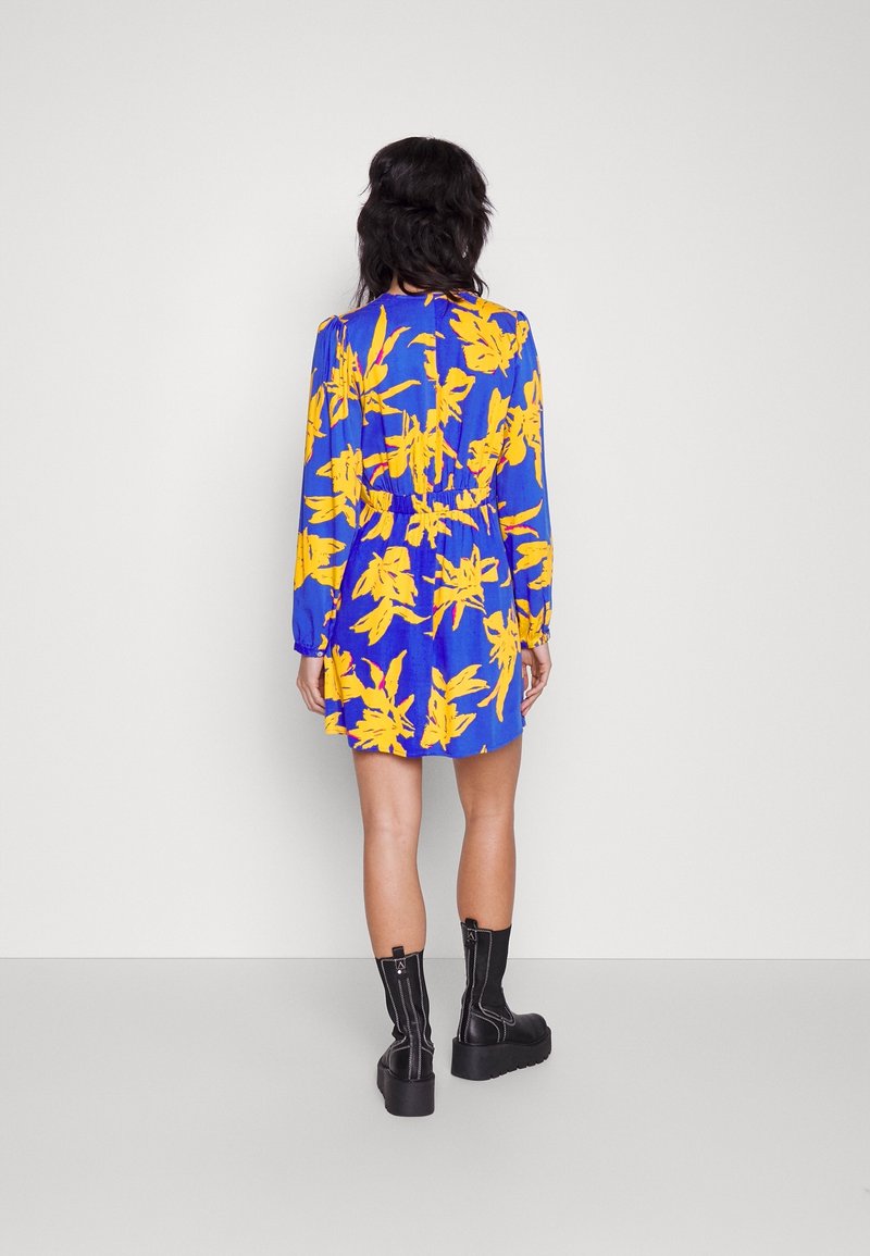 Robe bleue avec un grand motif floral jaune, manches longues, taille élastique et longueur au-dessus du genou. Associée à des bottines noires à plateforme.