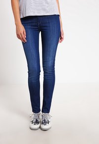 Jeans ajustados en denim azul oscuro con una textura suave. El modelo lleva zapatillas plateadas con acentos decorativos. Top de rayas simple.