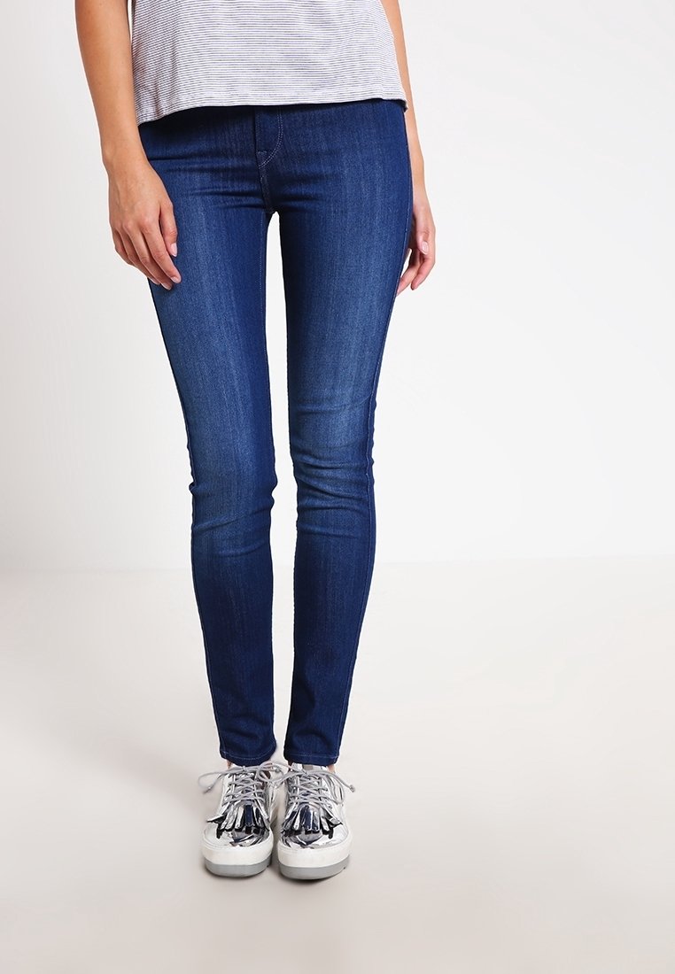 Jeans ajustados en denim azul oscuro con una textura suave. El modelo lleva zapatillas plateadas con acentos decorativos. Top de rayas simple.