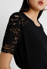 Černý top s krátkými rukávy vyrobený z propracované černé krajky, s kulatým výstřihem a hladkým tělem.