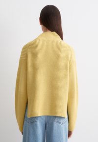 Żółty sweter dziany o prążkowanej strukturze, w oversize'owym kroju i z wysokim kołnierzem, połączony z jasnoniebieskimi dżinsami, widoczny z tyłu.