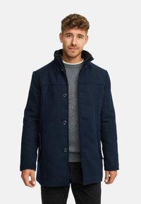 Marinblå ullblandad kappa med hög krage, knappstängning, sidofickor och en platt, strukturerad design. Bärs över en grå tröja.