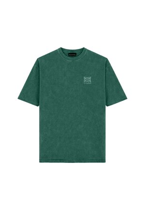 Man staat en draagt een groen oversized T-shirt met een klein logo, lichte denim shorts, witte sokken en zwart-witte sneakers tegen een effen achtergrond.