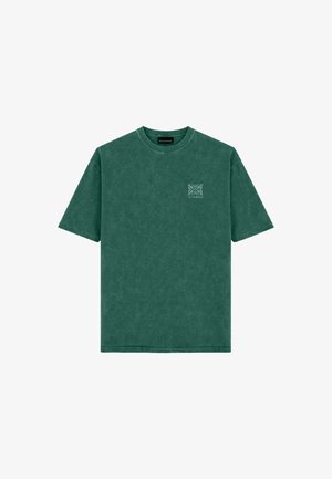 Mann steht und trägt ein grünes oversized T-Shirt mit kleinem Logo, helle Jeansshorts, weiße Socken und schwarz-weiße Turnschuhe vor einem einfarbigen Hintergrund.