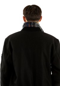 Muž viděný zezadu, oblečený v černé bundě a tmavém kostkovaném šále kolem krku, na bílém pozadí.