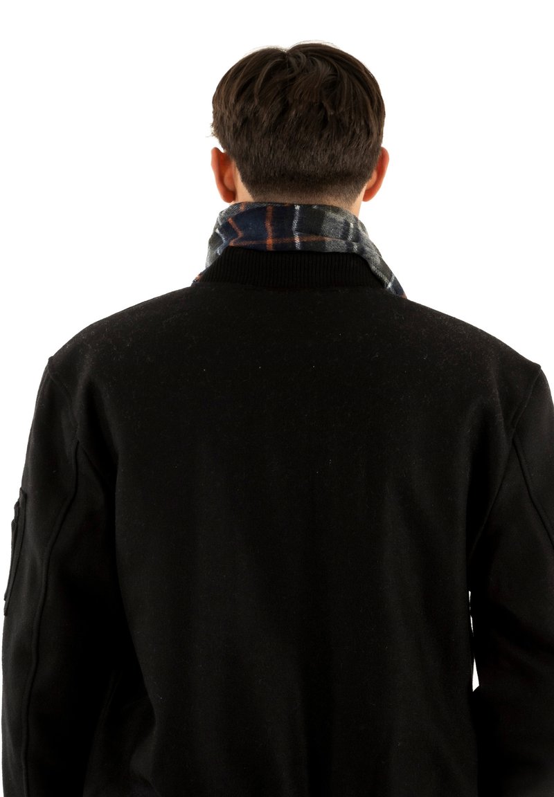 Muž viděný zezadu, oblečený v černé bundě a tmavém kostkovaném šále kolem krku, na bílém pozadí.