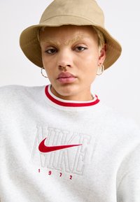 Sivi dukserice s rupčastim crvenim ovratnikom, sa vezenom "NIKE" logotipom u crvenoj i sivoj boji. Smeđa kape s obodom. Piercing na nosu i okrugle naušnice.