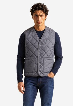 Mand med mørkt krøllet hår bærer en grå quiltet vest over en navyblå sweater og blå jeans, stående mod en ensfarvet hvid baggrund.