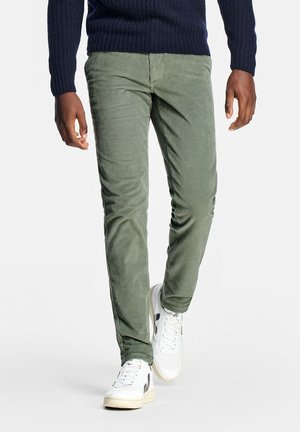 Homme marchant portant un pantalon en velours côtelé vert slim, un pull marine et des baskets blanches avec des accents noirs sur un fond blanc.