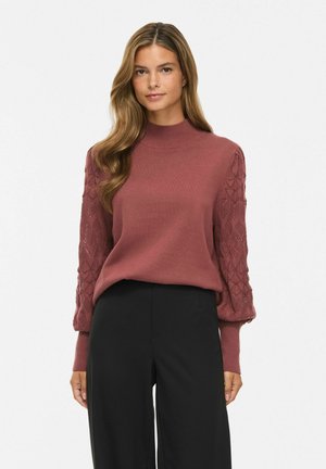 Modèle portant un pull rose poudré à col haut avec des manches à motifs et un pantalon large noir, debout sur un fond blanc uni.