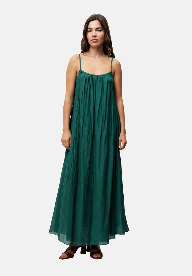 Maxi-jurk in diep groen, gemaakt van zachte, vloeiende stof. Beschikt over dunne bandjes en een geraffineerd lijfje. Platte schoenen getoond.