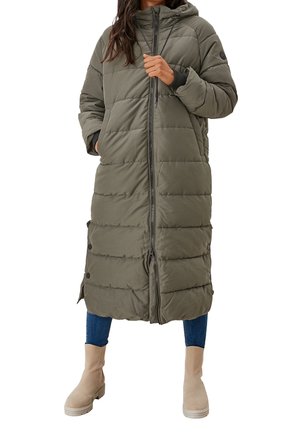 Personne portant un long manteau matelassé vert olive avec capuche, un jean bleu et des bottines beige, debout avec une main dans la poche et l'autre sur la fermeture éclair.