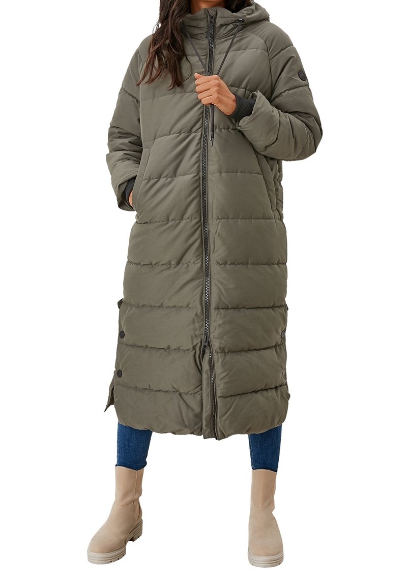 Personne portant un long manteau matelassé vert olive avec capuche, un jean bleu et des bottines beige, debout avec une main dans la poche et l'autre sur la fermeture éclair.