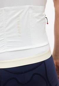 Κοντινό πλάνο ενός ατόμου που φοράει λευκό ποδηλατικό μπλουζάκι με υφή και την ένδειξη "Castelli" καθώς και μπλε σκουρόχρωμα ποδηλατικά σορτς με επένδυση.